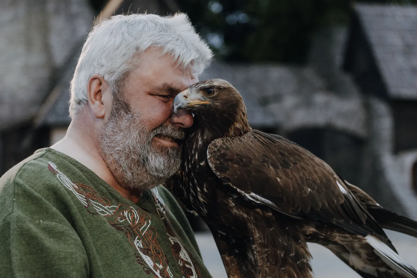 Ein Mann mit grauem Bart umarmt einen Adler in einer mittelalterlichen Kulisse.
