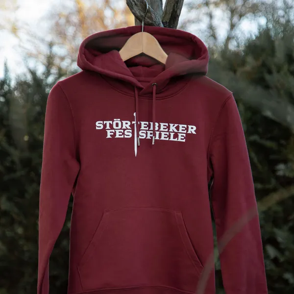 Roter Hoodie mit Aufdruck "Störtebeker Festspiele", aufgehängt vor grünen Büschen.