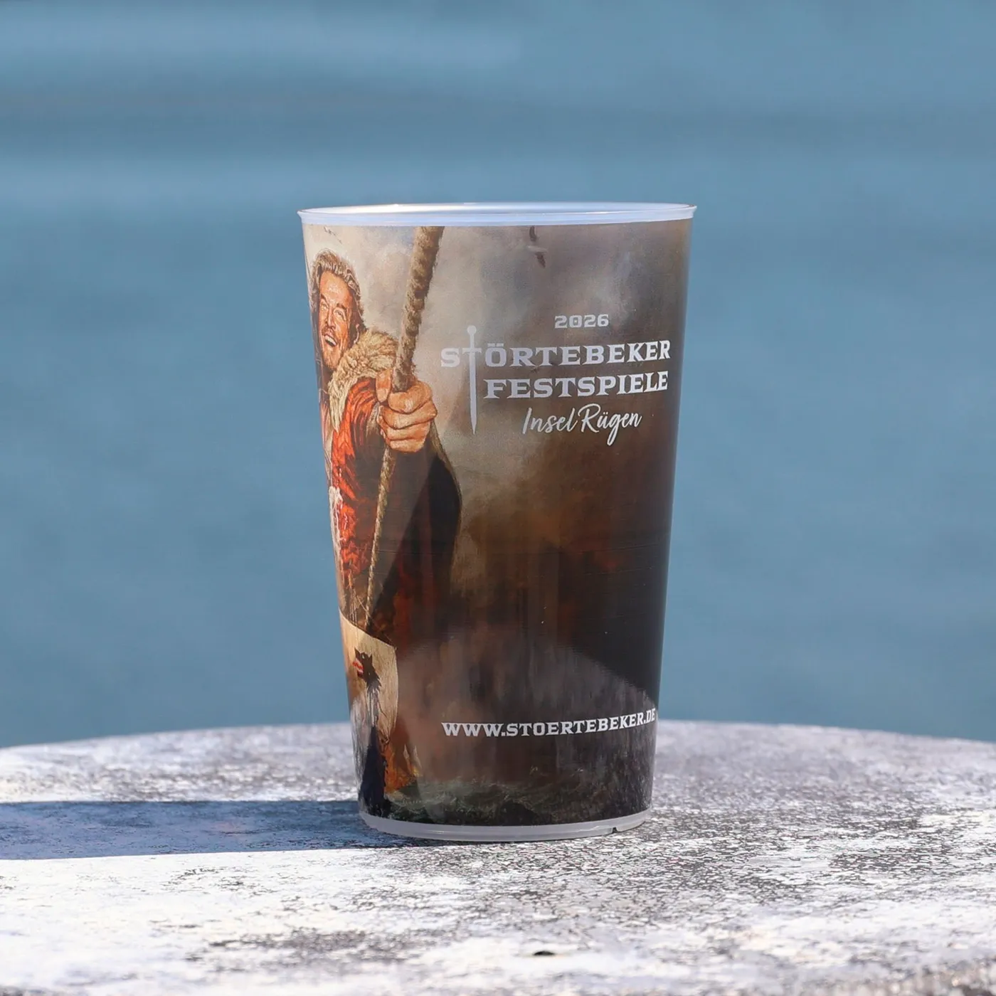 Becher mit Motiv der Störtebeker Festspiele, Hintergrund zeigt Wasser und Himmel.