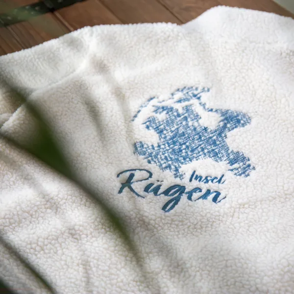 Weiße Fleecejacke mit blauem Aufdruck "Insel Rügen" auf einem Holztisch.