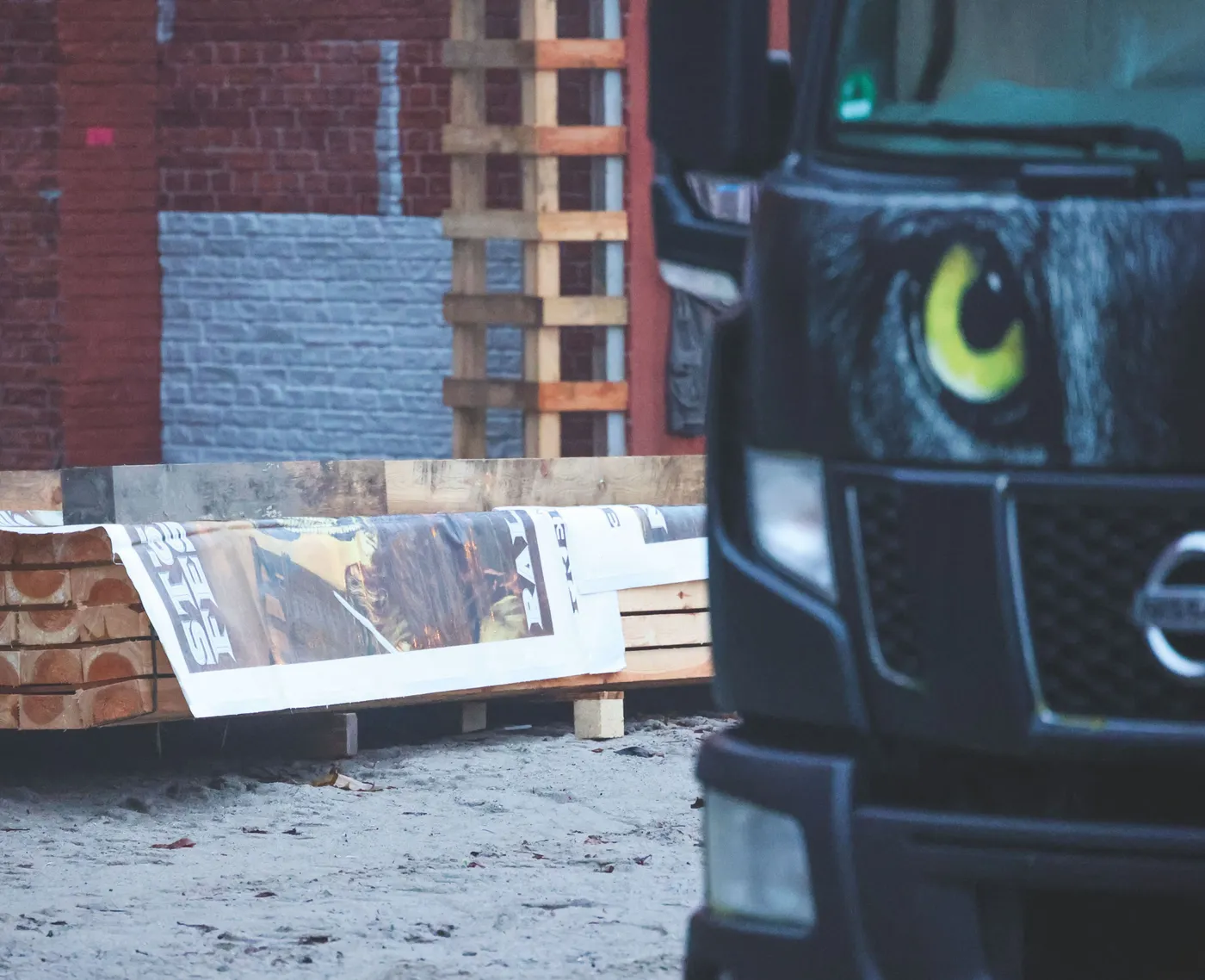 Holzstapel mit Plakaten im Hintergrund, im Vordergrund ein Fahrzeug mit auffälligem Design.