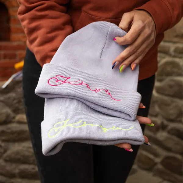 Zwei graue Mützen mit bunten Schriftzügen werden von einer Person gehalten.