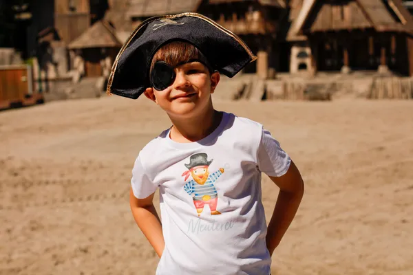 Ein Junge im Piratenkostüm mit Augenklappe steht lächelnd auf einem Sandplatz.
